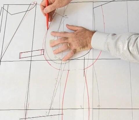 Mani di un modellista che disegna un cartamodello su carta tecnica, tracciando linee e curve con una matita rossa per la creazione di un capo di abbigliamento